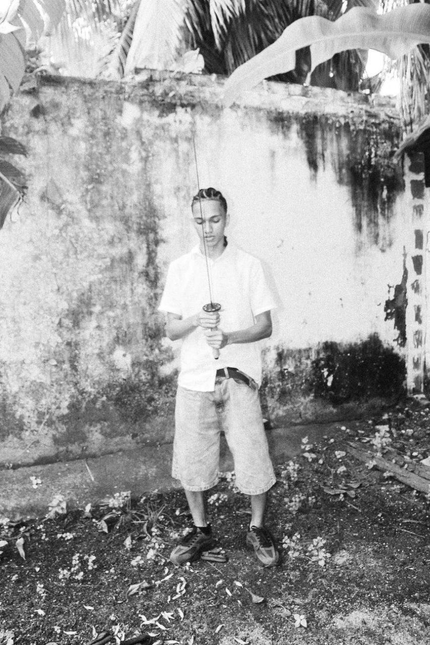 Fotografia em preto e branco de Rakim em pé diante de um muro desgastado, segurando uma katana, cercado de folhagem tropical